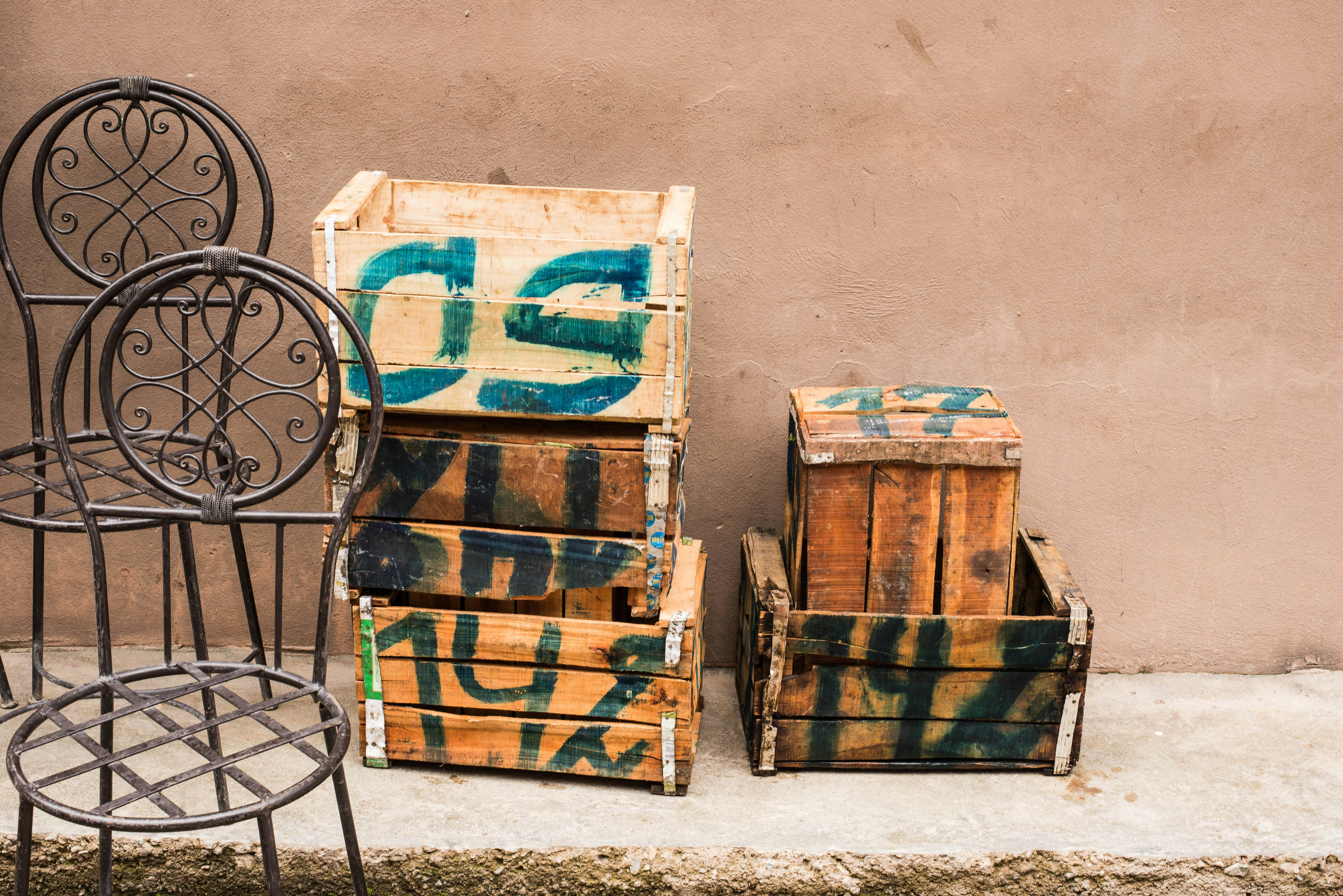 Several wooden crates with green paint markings are stacked against a plain beige wall. Two metal chairs with ornate, circular backrests are placed next to the crates on a concrete surface.
