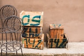 Several wooden crates with green paint markings are stacked against a plain beige wall. Two metal chairs with ornate, circular backrests are placed next to the crates on a concrete surface.