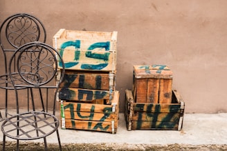 Several wooden crates with green paint markings are stacked against a plain beige wall. Two metal chairs with ornate, circular backrests are placed next to the crates on a concrete surface.