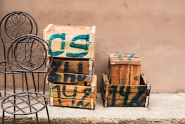 Several wooden crates with green paint markings are stacked against a plain beige wall. Two metal chairs with ornate, circular backrests are placed next to the crates on a concrete surface.