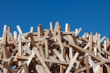 A chaotic arrangement of wooden planks intertwined, creating a complex, three-dimensional structure against a clear blue sky.