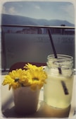 A mason jar filled with lemonade and a black straw is placed next to a white pot containing bright yellow flowers. The scene is set on a table with a backdrop of a cityscape and mountains in the distance, framed by a clear blue sky.