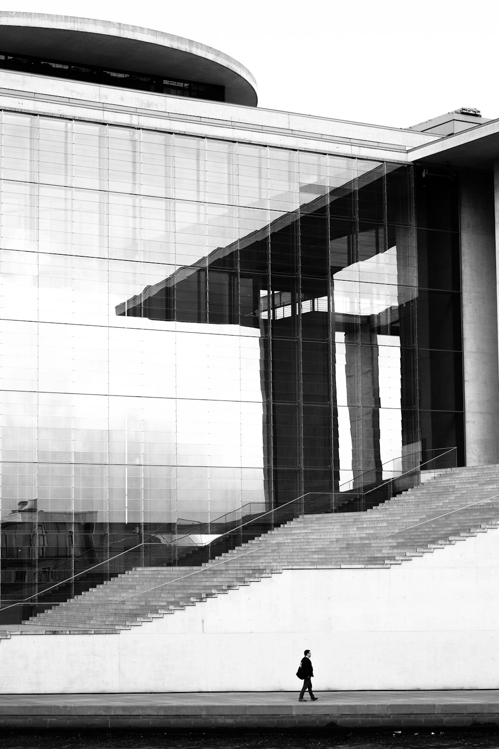 Foto en escala de grises del hombre caminando sobre el pavimento de hormigón al lado de un edificio alto
