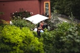 A small group of people gather under a white canopy in a lush garden next to a building with a brown tiled roof and large window. The scene is surrounded by dense green foliage, creating an intimate and natural setting.