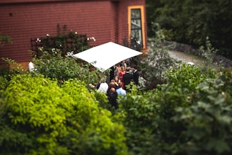 A small group of people gather under a white canopy in a lush garden next to a building with a brown tiled roof and large window. The scene is surrounded by dense green foliage, creating an intimate and natural setting.