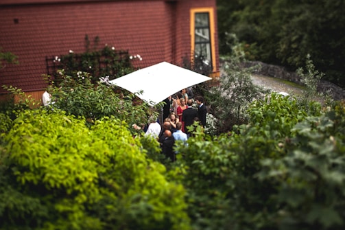 A small group of people gather under a white canopy in a lush garden next to a building with a brown tiled roof and large window. The scene is surrounded by dense green foliage, creating an intimate and natural setting.