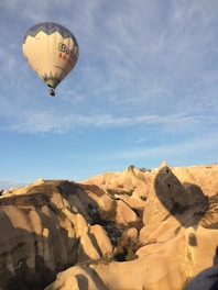 A hot air balloon glides gracefully over a unique and rugged landscape of rocky formations. The balloon features a pattern with a mixture of neutral and dark colors, contrasted against the pale, sunlit rocks below. A vast blue sky stretches above, speckled with wispy clouds.