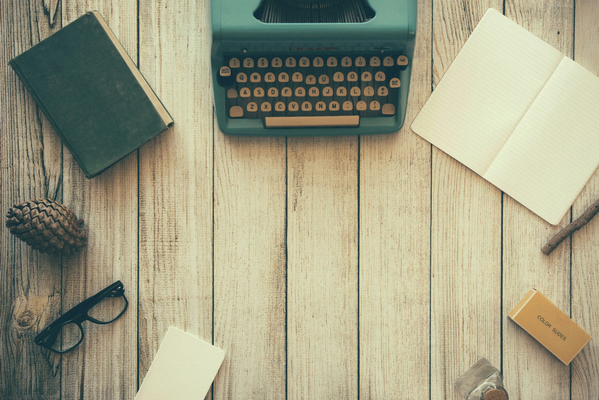 Wooden desk with a typewriter, and open blank notebook and a few other items that, perhaps, induce creativity.