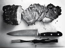 A black and white photo displays a sliced roast on a cutting surface. Several slices reveal the tender interior of the meat and a crusty outer layer. Below the slices, a sharp kitchen knife and a two-pronged fork rest parallel to each other.