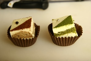 Two small square cakes placed in brown paper liners are displayed, each with a geometric topping. The left cake has a chocolate and cream patterned top with a small chocolate adornment, while the right cake features a matcha green tea and cream design accented with a metallic pearl.