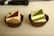Two small square cakes placed in brown paper liners are displayed, each with a geometric topping. The left cake has a chocolate and cream patterned top with a small chocolate adornment, while the right cake features a matcha green tea and cream design accented with a metallic pearl.