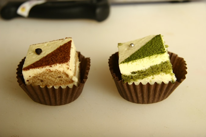 Two small square cakes placed in brown paper liners are displayed, each with a geometric topping. The left cake has a chocolate and cream patterned top with a small chocolate adornment, while the right cake features a matcha green tea and cream design accented with a metallic pearl.