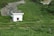 A small white building with a green tiled roof is situated amidst lush green grassy surroundings. The area is nestled in a peaceful, rural setting with a narrow dirt path leading to the structure. A small creek or stream runs adjacent to the building, bordered by rocks and greenery.