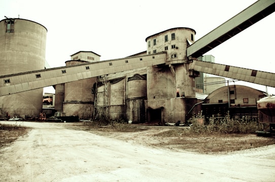 Industrial cement factory with large silos and conveyor belts surrounded by rugged terrain.