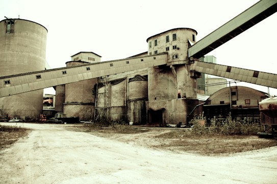 Industrial site with large concrete silos and elevated conveyor belts crossing over them. The ground is barren and there are signs of neglect with overgrown weeds. The structure appears to be worn and aged, with visible rust and dirt.