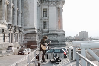 A musician with curly hair stands on an outdoor platform beside a historic stone building, playing a guitar. The scene overlooks a cityscape with buildings visible in the background and a few parked cars nearby. The majestic structure features tall columns and intricate architectural details.