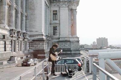 A musician with curly hair stands on an outdoor platform beside a historic stone building, playing a guitar. The scene overlooks a cityscape with buildings visible in the background and a few parked cars nearby. The majestic structure features tall columns and intricate architectural details.