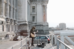 A musician with curly hair stands on an outdoor platform beside a historic stone building, playing a guitar. The scene overlooks a cityscape with buildings visible in the background and a few parked cars nearby. The majestic structure features tall columns and intricate architectural details.