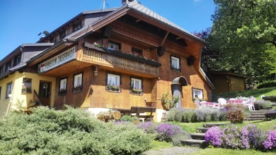 A rustic mountain lodge with a wooden exterior is surrounded by lush greenery and colorful flowers. The building features several small windows adorned with flower boxes, a balcony, and a shingled roof. Outdoor seating with white chairs and tables is visible on the lawn, which is beautifully landscaped with purple and pink flowering plants and shrubs.
