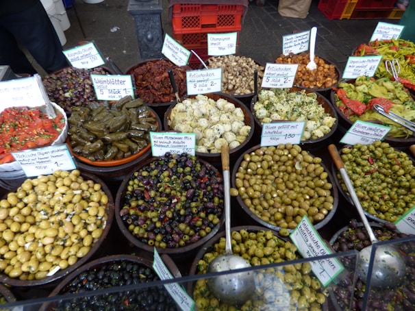 A variety of marinated olives and other Mediterranean delicacies are displayed in large bowls, each labeled with a sign. The selection includes stuffed grape leaves, sun-dried tomatoes, artichokes, and various types of seasoned olives. Each dish is artfully arranged with serving spoons, emphasizing the rich colors and textures of the food.
