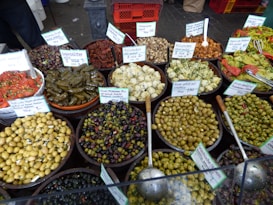 A variety of marinated olives and other Mediterranean delicacies are displayed in large bowls, each labeled with a sign. The selection includes stuffed grape leaves, sun-dried tomatoes, artichokes, and various types of seasoned olives. Each dish is artfully arranged with serving spoons, emphasizing the rich colors and textures of the food.