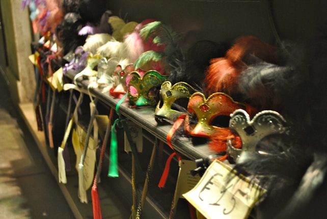 A collection of colorful masquerade masks displayed on a shelf, each adorned with feathers, glitter, and intricate designs. Price tags are visible, indicating they are for sale. The setting appears to be dimly lit, creating a mysterious and festive atmosphere.