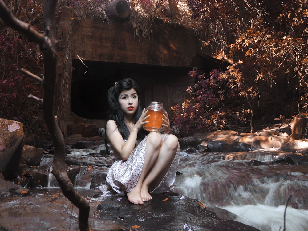 woman carrying glass jar while sitting on boulder beside flowing river behind brown bridge under shade of tree at daytime