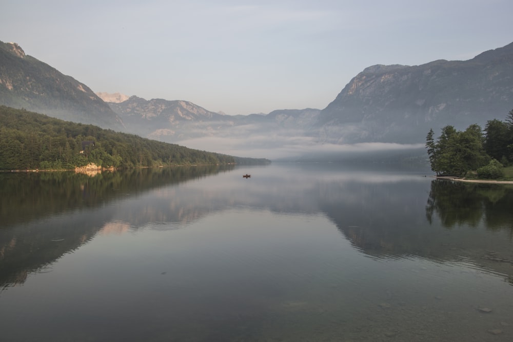 body of water near mountain ranges with green vegetation covered with fog during daytime