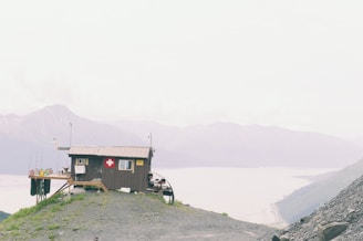 A small wooden cabin with a red cross emblem is perched on a rocky hilltop, overlooking expansive mountains and a river in the background. The sky is overcast, creating a muted atmosphere.