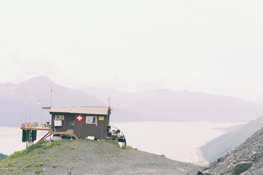 A small wooden cabin with a red cross emblem is perched on a rocky hilltop, overlooking expansive mountains and a river in the background. The sky is overcast, creating a muted atmosphere.