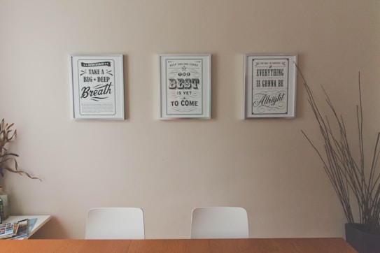 Three framed motivational posters are displayed on a beige wall above a wooden table and two white chairs. The left poster reads 'Take a Big Deep Breath', the center one says 'The Best is Yet to Come', and the right one states 'Everything is Gonna Be Alright'. A few magazines or brochures are visible on the corner of the table. Tall, decorative plant stems are placed in a vase on the right side.