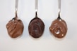 Three spoons are arranged side by side, each holding a different type of thick, chocolatey spread. The textures of the spreads vary from smooth to slightly granular.