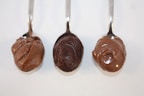 Three spoons are arranged side by side, each holding a different type of thick, chocolatey spread. The textures of the spreads vary from smooth to slightly granular.