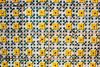 A wall covered with ornate tiles featuring a repeated pattern of yellow flowers with green leaves on a white background. The tiles have a weathered texture, suggesting age or outdoor exposure.