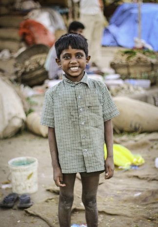 A young boy with a big smile stands in a busy, market-like setting. Wearing a checkered shirt, he appears happy and confident amidst the blurred background showing baskets, sacks, and fabrics.