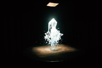 A dynamic splash of water is suspended in mid-air, illuminated by a spotlight from above. The background is dark, enhancing the glowing effect of the water.