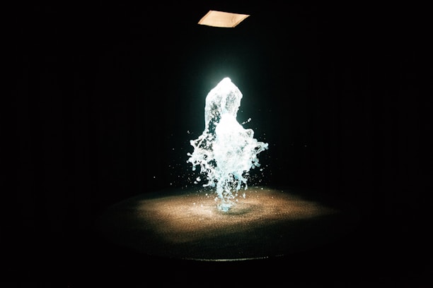 A dynamic splash of water is suspended in mid-air, illuminated by a spotlight from above. The background is dark, enhancing the glowing effect of the water.