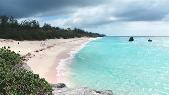 A serene beach with light pink sand stretches along the coastline, meeting turquoise blue ocean waters. The sky is partly cloudy, casting a soft shadow over the landscape. Rocks and sparse beachgoers are visible in the distance, while lush green vegetation borders the sand.