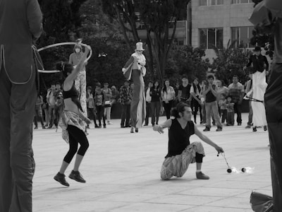 A street performance scene with performers dressed in various costumes, including one on stilts and another juggling fire. A crowd of onlookers gathers in the background, observing the entertainment. The setting appears to be an outdoor public space lined with trees and buildings.