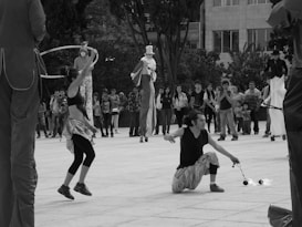 A street performance scene with performers dressed in various costumes, including one on stilts and another juggling fire. A crowd of onlookers gathers in the background, observing the entertainment. The setting appears to be an outdoor public space lined with trees and buildings.