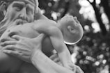 A monochrome photograph capturing a close-up of a detailed sculpture featuring a bearded man holding an infant. The man's expression appears contemplative, and the infant is depicted looking upwards with a serene expression. The background is blurred with circular bokeh lights.