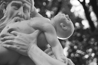 A monochrome photograph capturing a close-up of a detailed sculpture featuring a bearded man holding an infant. The man's expression appears contemplative, and the infant is depicted looking upwards with a serene expression. The background is blurred with circular bokeh lights.