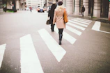A woman walking through a sunlit city street wearing a beige trench coat and light scarf.