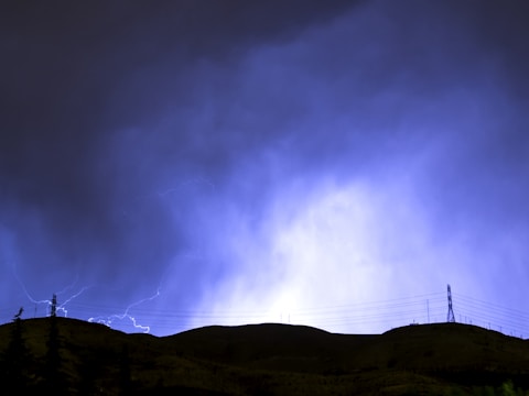 A dramatic scene with a dark silhouette of hills and trees under a stormy sky illuminated by bright blue lightning. Power lines and a pylon are visible against the contrasting light of the storm.