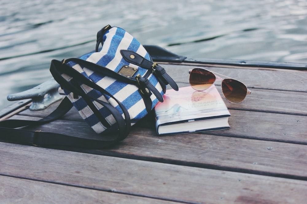Blau-weiß gestreifte Umhängetasche in der Nähe von Buch und Sonnenbrille auf dem Seedock während des Tages