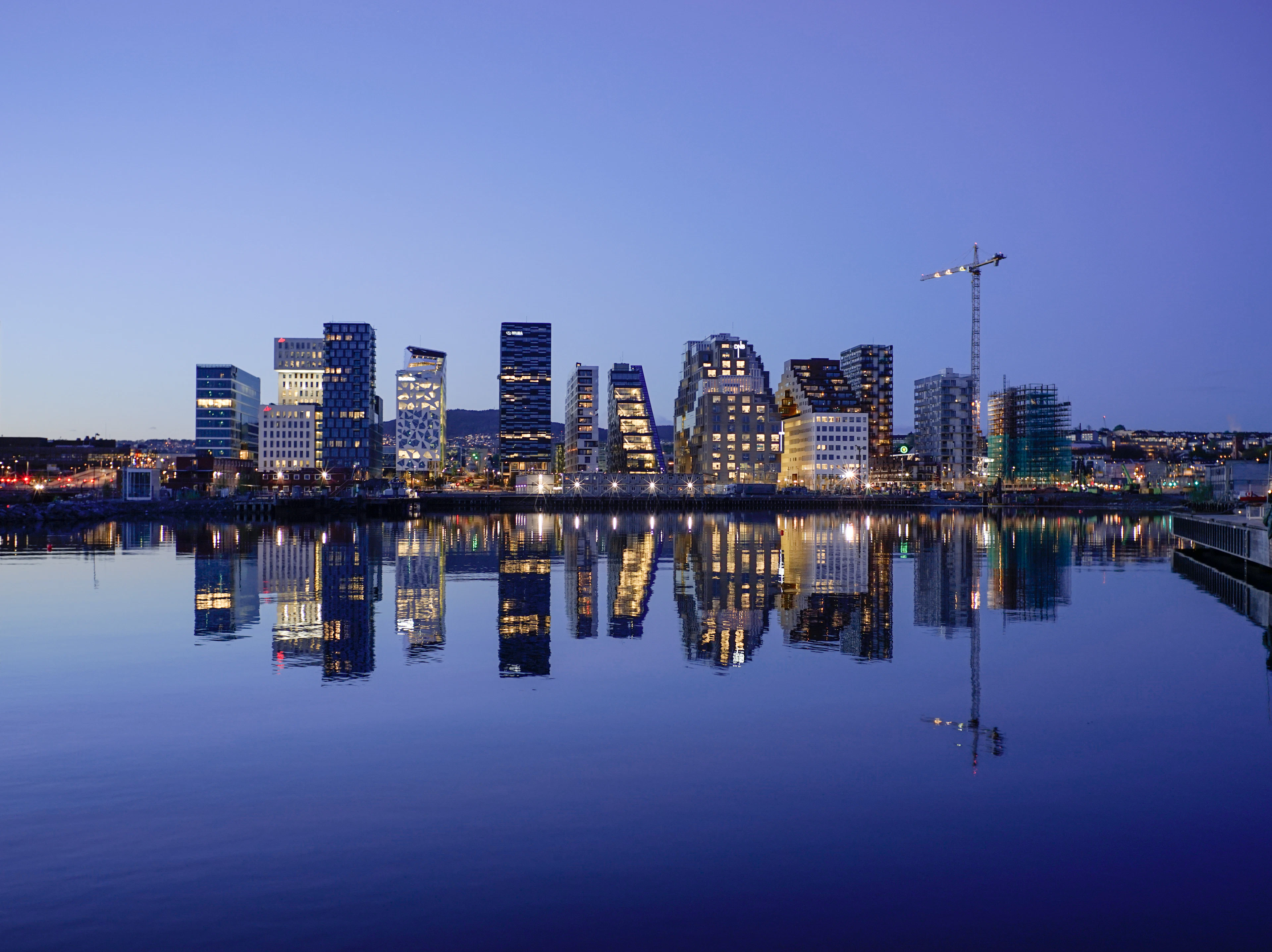 The Barcode towers of Oslo's Bjorvika waterfront at blue hour, their lit windows and angular glass facades mirrored almost perfectly in the flat water of the harbour under a clear violet sky