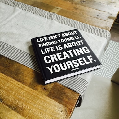 A black hardcover book with white text on the cover is placed on a wooden table. A light gray table runner with striped edges lies underneath the book. The book's cover reads, 'Life isn't about finding yourself. Life is about creating yourself.' The image also features the corner of a patterned rug in the background.