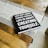 A black hardcover book with white text on the cover is placed on a wooden table. A light gray table runner with striped edges lies underneath the book. The book's cover reads, 'Life isn't about finding yourself. Life is about creating yourself.' The image also features the corner of a patterned rug in the background.