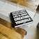 A black hardcover book with white text on the cover is placed on a wooden table. A light gray table runner with striped edges lies underneath the book. The book's cover reads, 'Life isn't about finding yourself. Life is about creating yourself.' The image also features the corner of a patterned rug in the background.