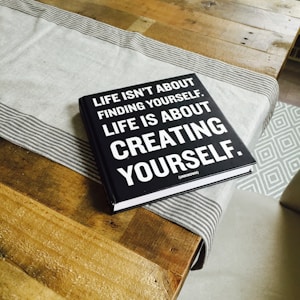 A black hardcover book with white text on the cover is placed on a wooden table. A light gray table runner with striped edges lies underneath the book. The book's cover reads, 'Life isn't about finding yourself. Life is about creating yourself.' The image also features the corner of a patterned rug in the background.